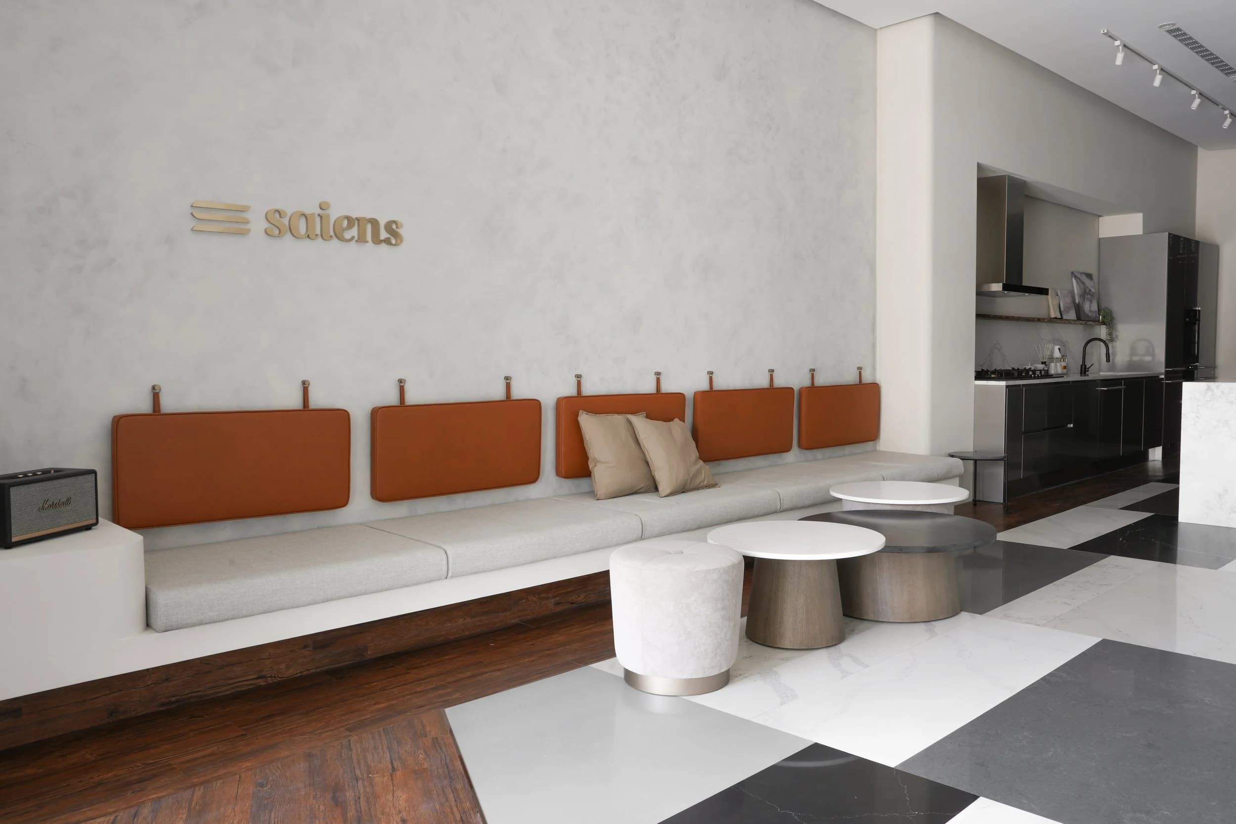 Modern minimalist waiting area with a long beige sofa, orange headboard-style cushions, a small black Marshall speaker, and a set of minimalist white and gray tiered coffee tables.